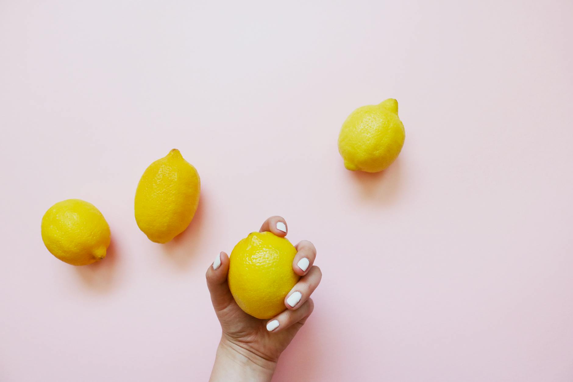 Una mano sostiene un limón fresco sobre un fondo rosa suave, rodeado de tres limones adicionales