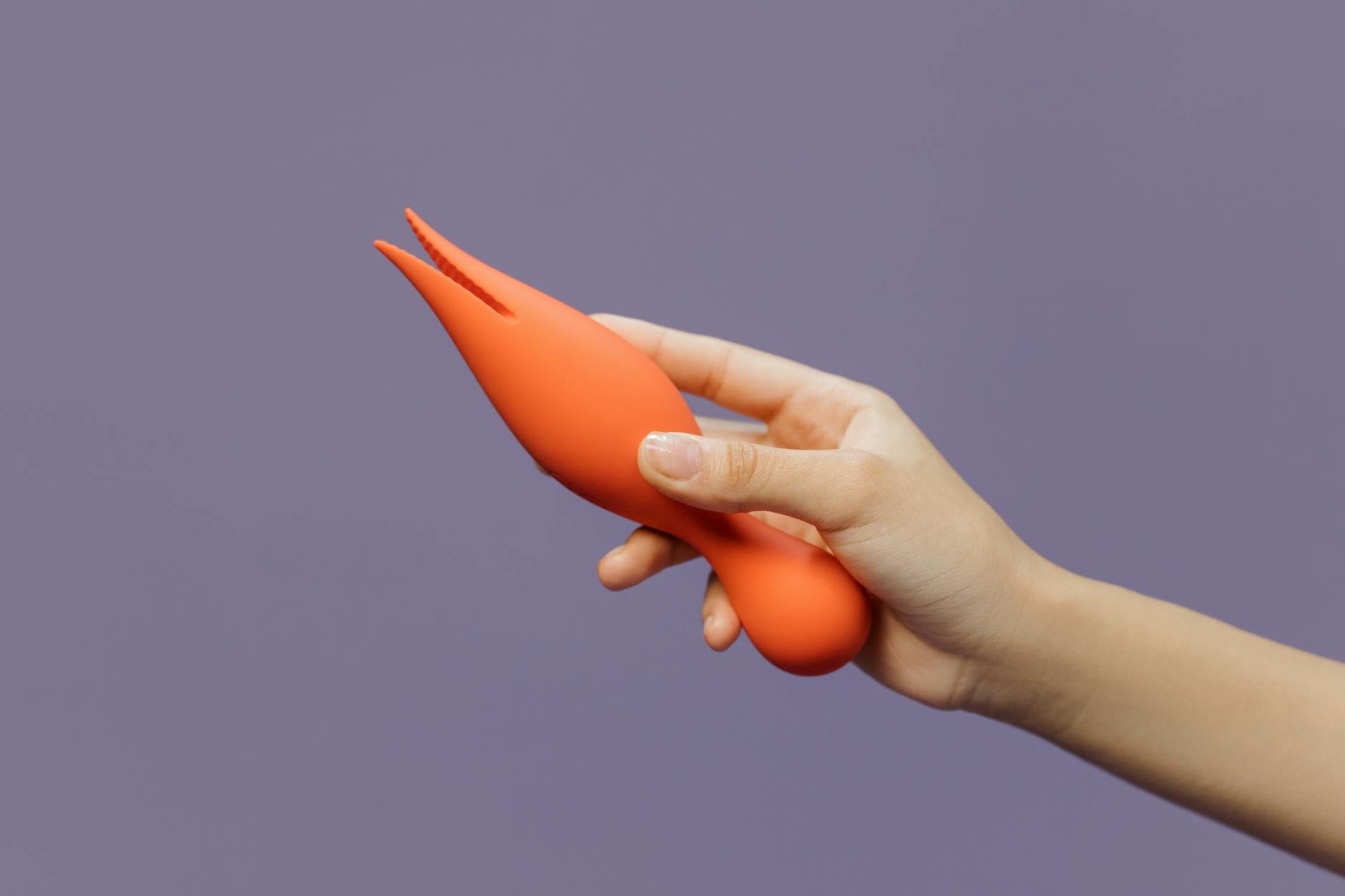 Close-up of a hand holding an orange vibrator against a minimalistic purple backdrop, showcasing modern sensuality