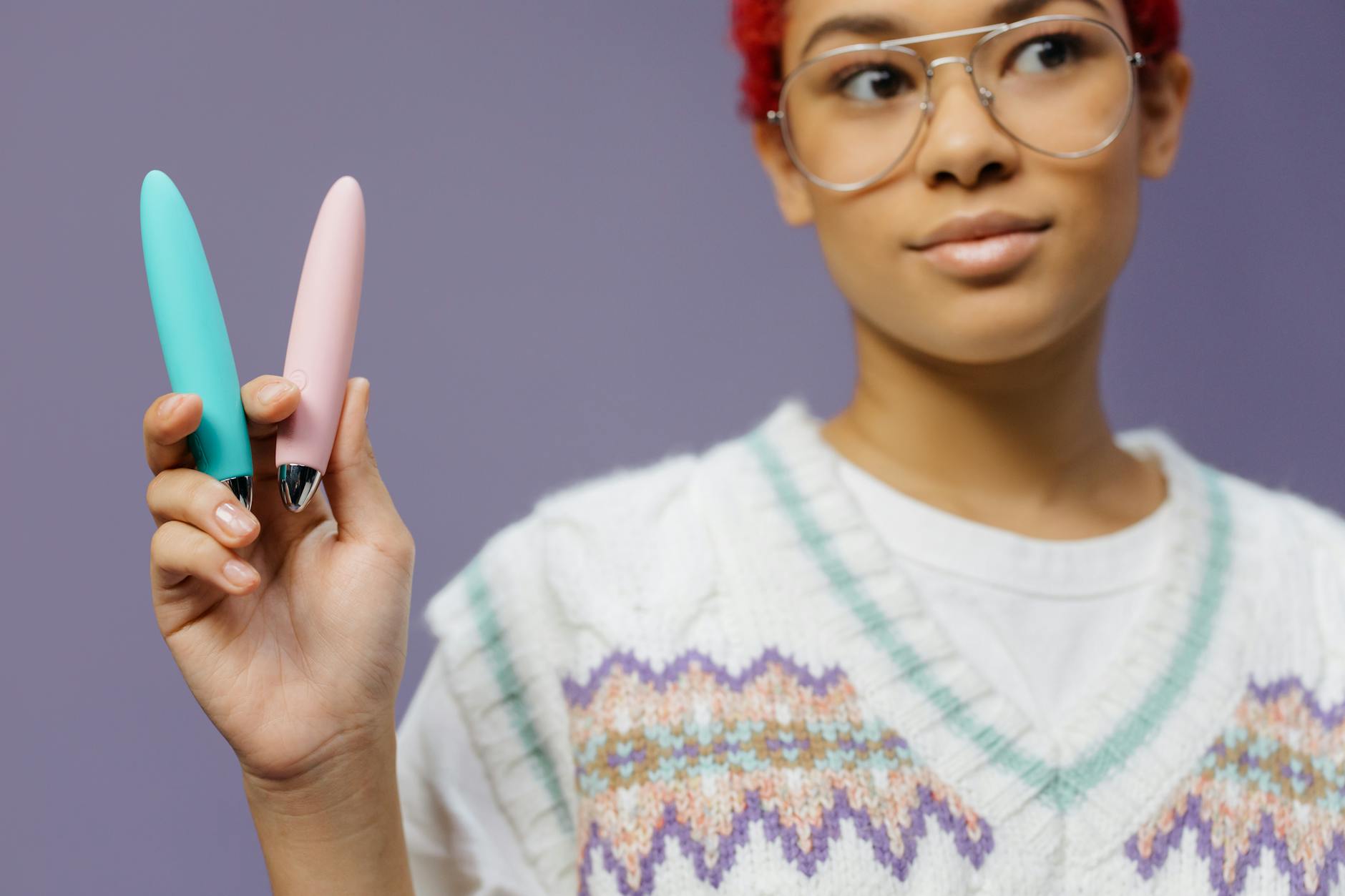 Mujer joven con gafas sosteniendo dos vibradores coloridos contra un fondo morado